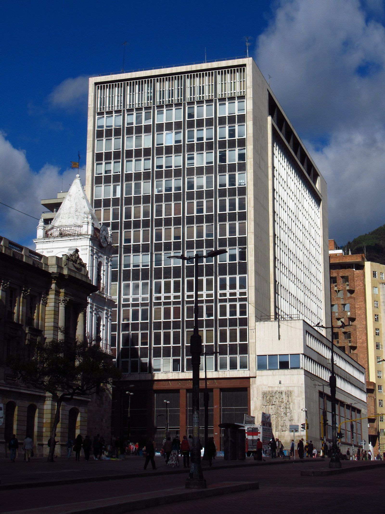 Torre de oficinas de vidrio y concreto del edificio del Banco de la República en Bogotá, con una iglesia colonial blanca a la izquierda